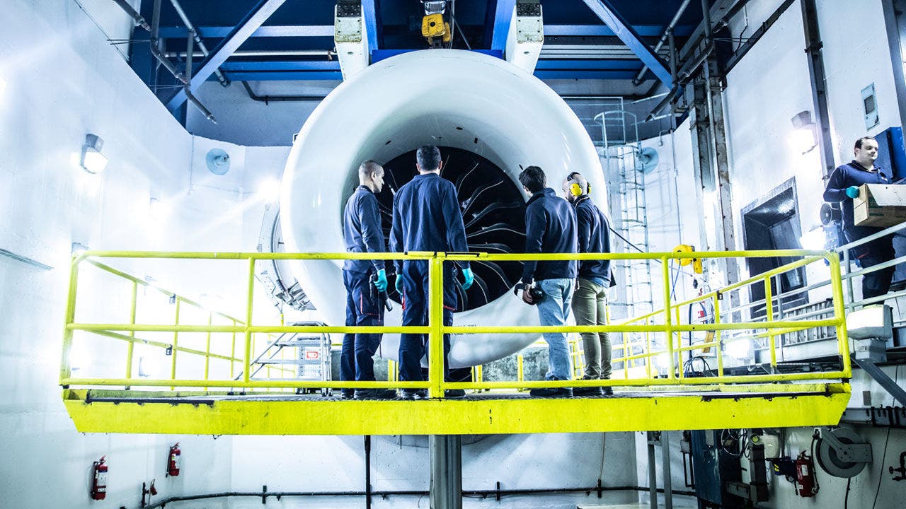 Four TAP maintenance workers standing on a platform in front of a large jet engine. On the right a worker carries a package.