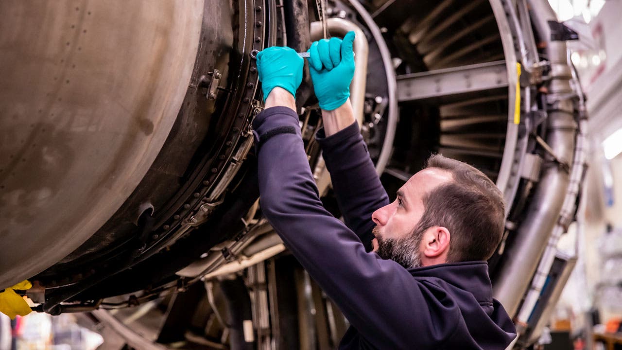 TAP Maintenance technician working on a jet engine during maintenance. He is wearing a dark blue uniform and teal protective gloves.