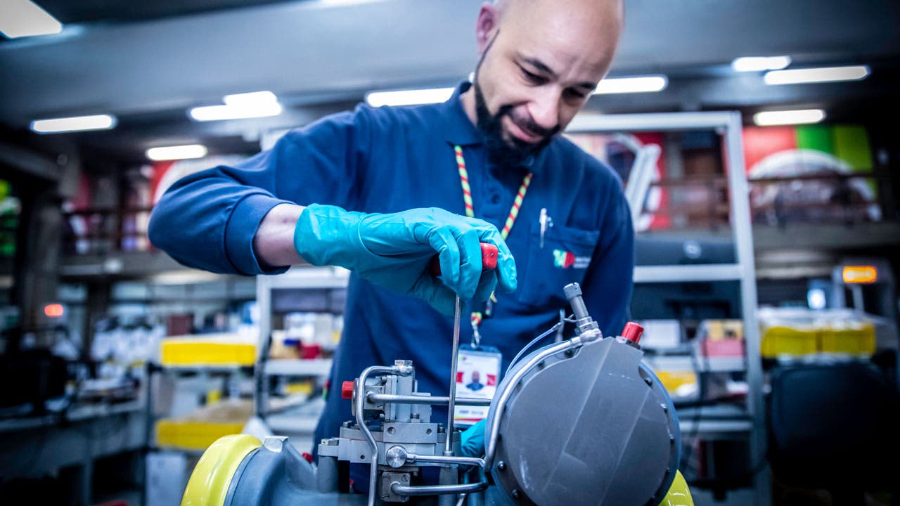 TAP Maintenance worker working on a mechanical component in a industrial setting. he technician is wearing teal-colored protective gloves and a blue uniform, with a visible TAP ID badge and a lanyard around his neck. He appears to be using a screwdriver.