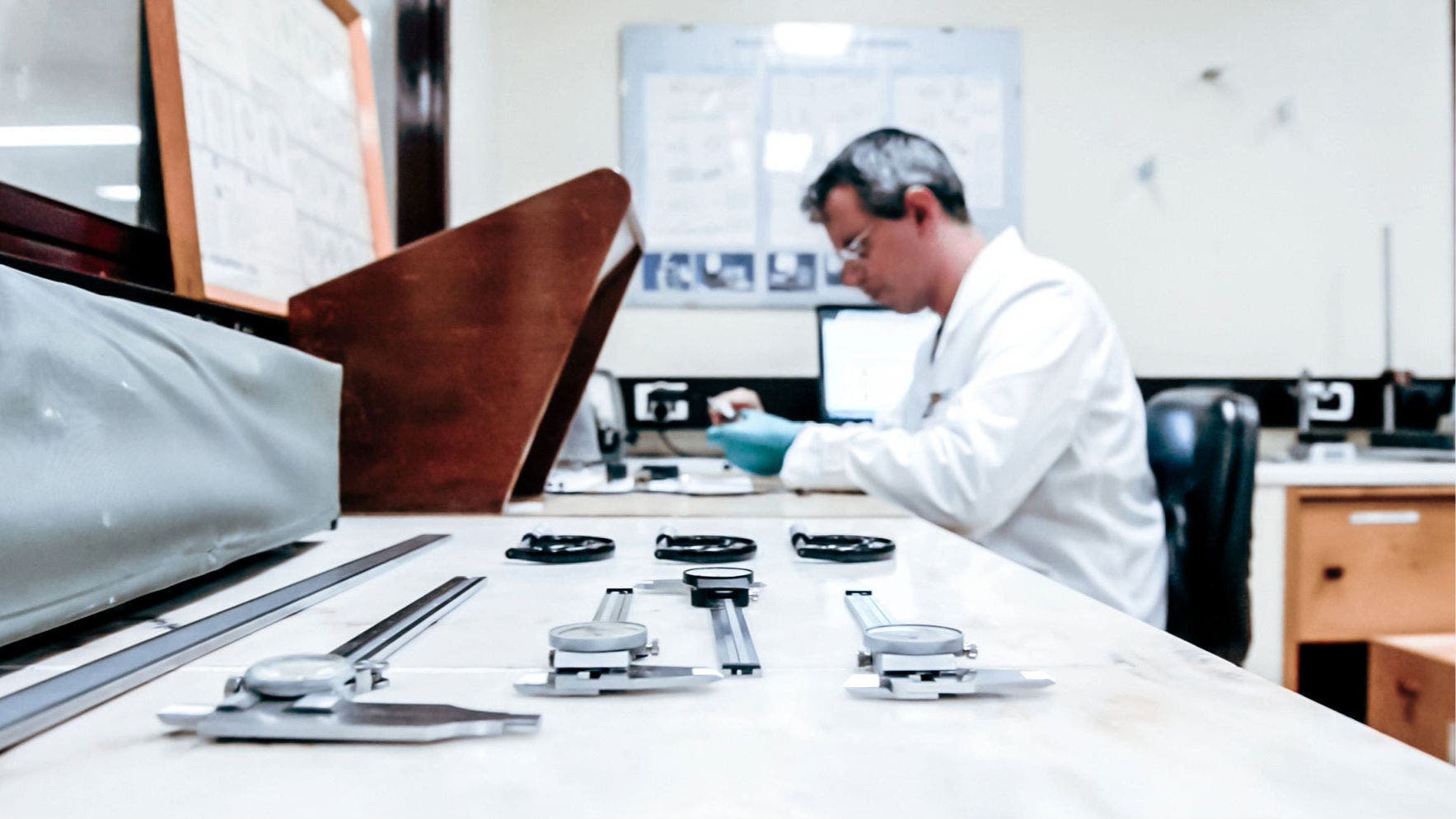Laboratory environment with a technician in a white lab coat and blue gloves focused on a task in the background. In the foreground, there are several dial indicators and calipers laid out on a workbench.