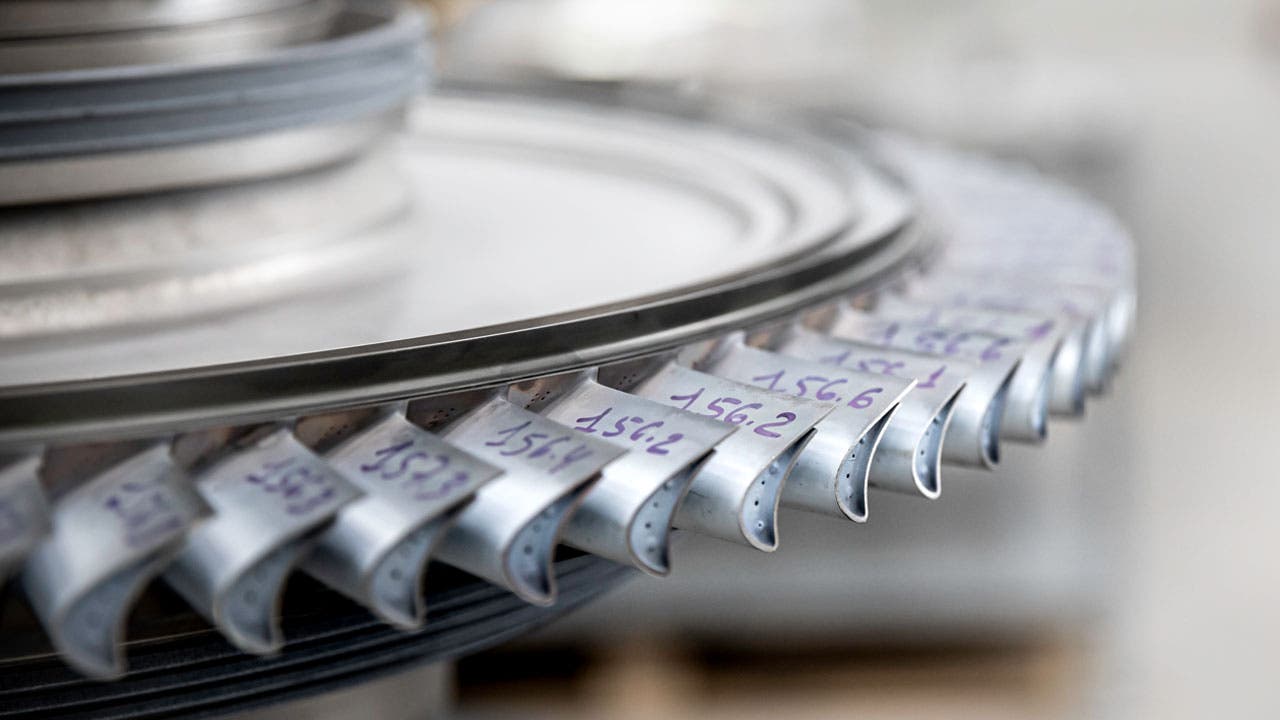 Close-up of a turbine or compressor disk from a jet engine. The component features several precision-engineered blades arranged radially around the disk. Each blade has a number written on it.