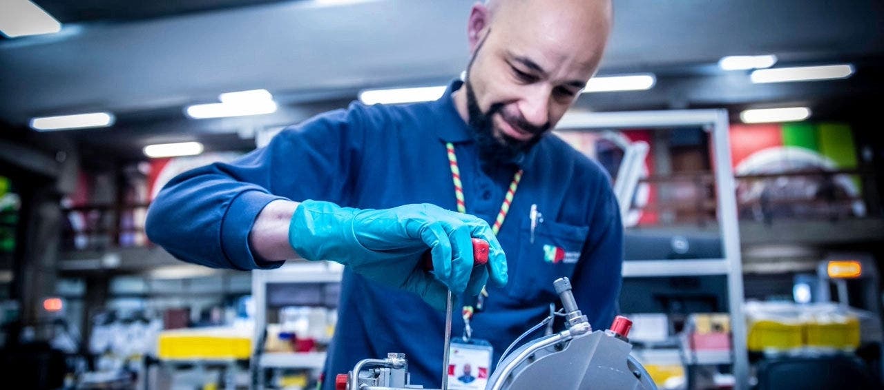 TAP Maintenance worker working on a mechanical component in a industrial setting. he technician is wearing teal-colored protective gloves and a blue uniform, with a visible TAP ID badge and a lanyard around his neck. He appears to be using a screwdriver.