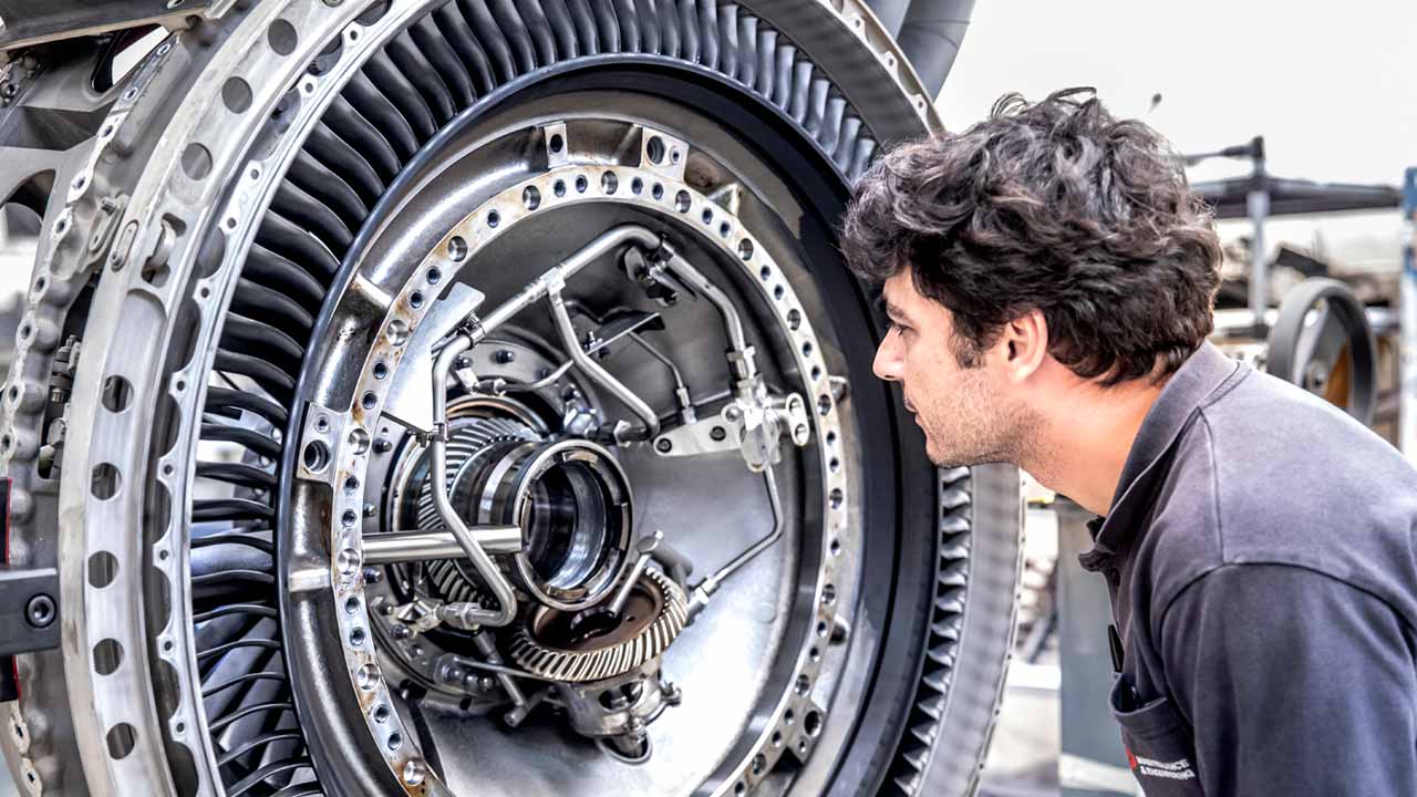 A TAP Maintenance worker inspects internal components of a jet engine. The engine is partially disassembled, revealing detailed parts such as turbine blades, gears, piping, and structural elements.