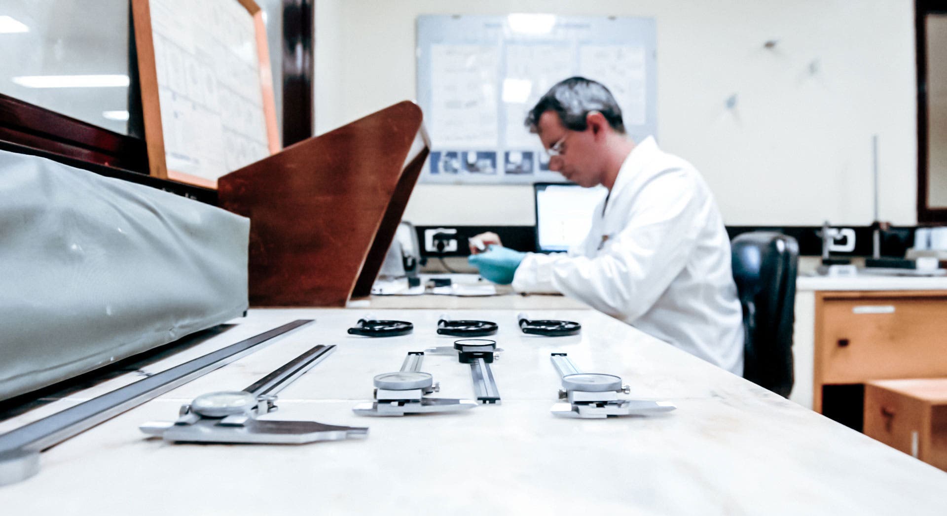 Laboratory environment with a technician in a white lab coat and blue gloves focused on a task in the background. In the foreground, there are several dial indicators and calipers laid out on a workbench.
