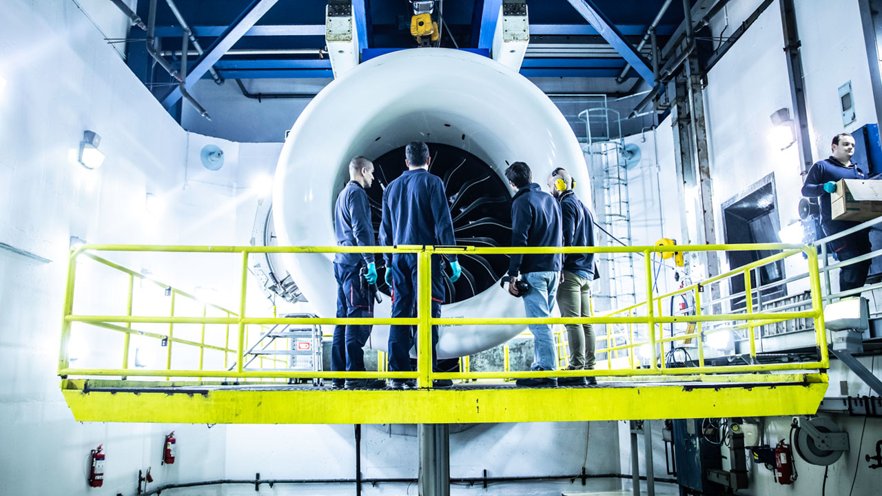 Four TAP maintenance workers standing on a platform in front of a large jet engine. On the right a worker carries a package.